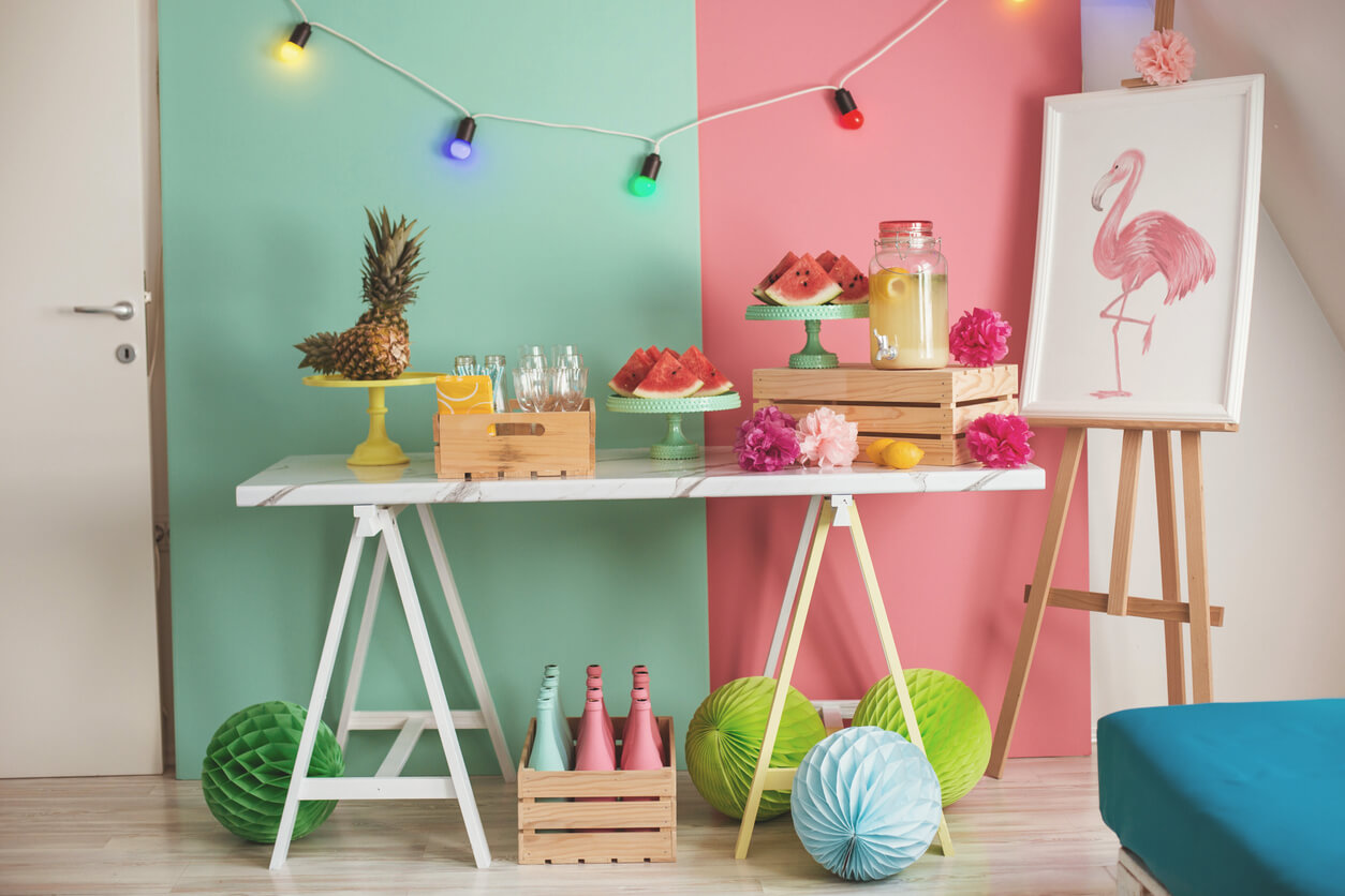 DIY party decoration ideas spread out among a colorful room, including a painting on an easel, fruits and lemonade on a table, and tissue paper poms on the floor